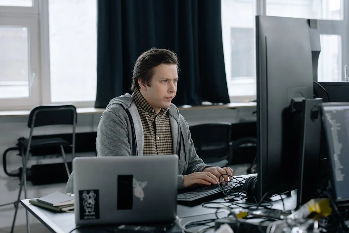 Man in gray hoodie working at a desk with dual monitors and a laptop in an office