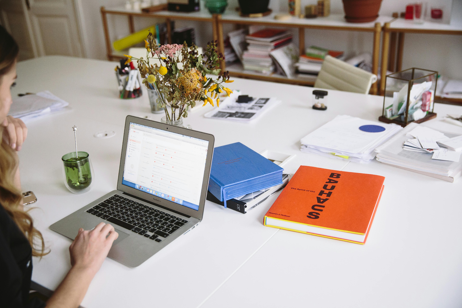Woman working on a laptop at a white desk with books and flowers.