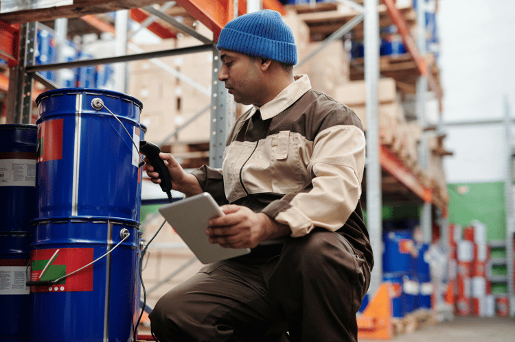 Warehouse worker scanning a blue drum's barcode with a handheld scanner while holding a tablet.